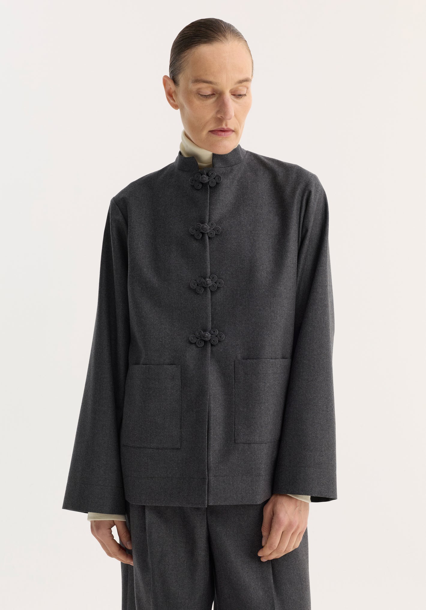 Person wearing Róhe 'Marie' mandarin jacket in charcoal-gray with pankou closures, embroidered edges, and patch pockets, paired with matching trousers.