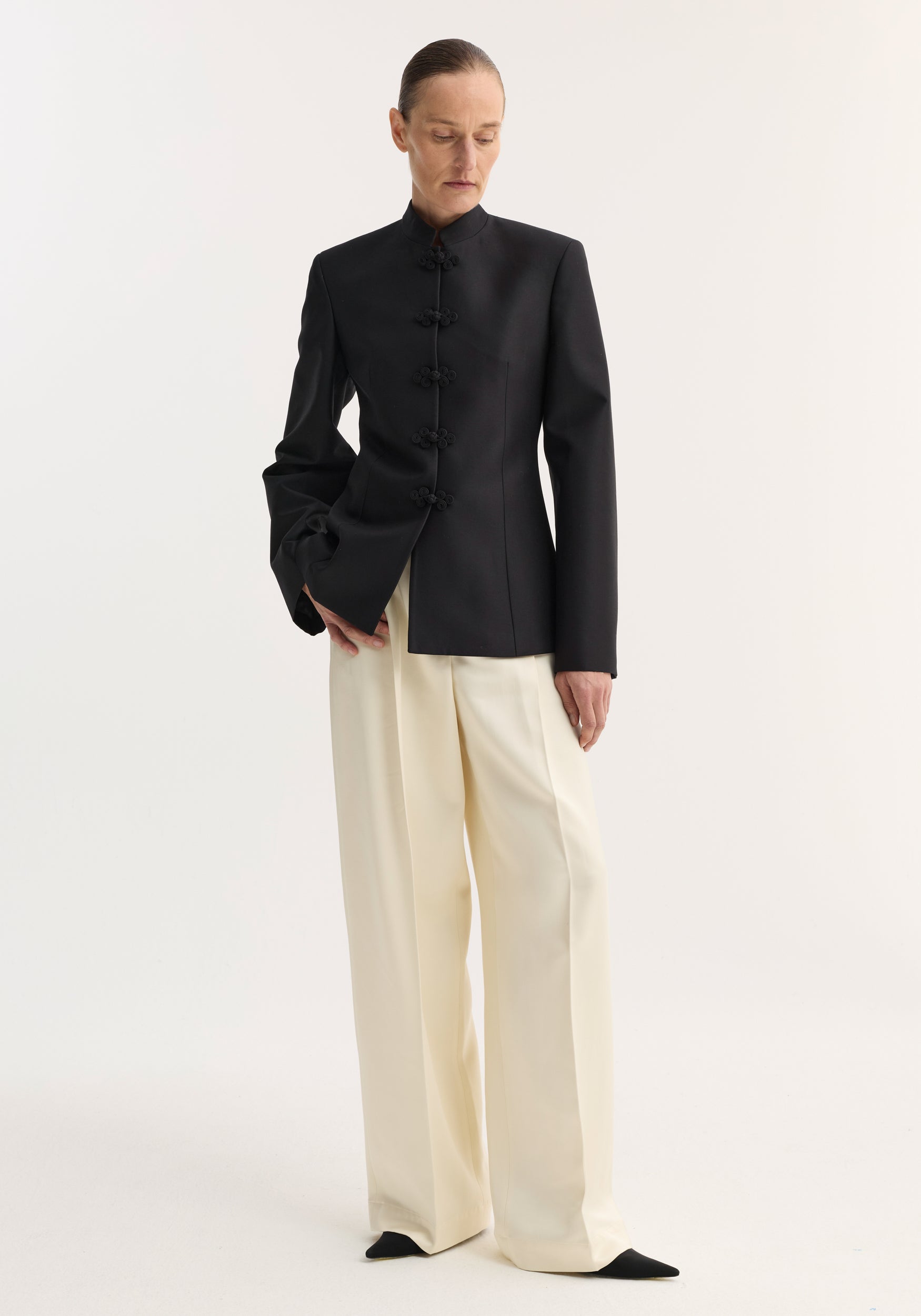 Model in a sculptural mandarin blazer, featuring pankou closures, tailored fit, and refined shoulders, paired with high-waisted, wide-leg trousers and pointed-toe shoes.