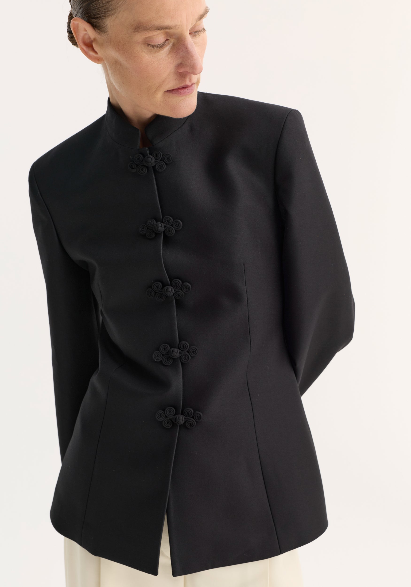 Sculptural mandarin blazer in black with pankou closures, worn by a woman, showcasing a fitted waist and refined shoulders. Made from a wool-silk blend.