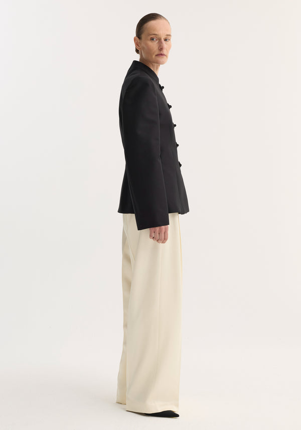 Sculptural mandarin blazer in black featuring pankou closures, tailored fit, and refined shoulders. Model wears it with high-waisted, wide-leg trousers.