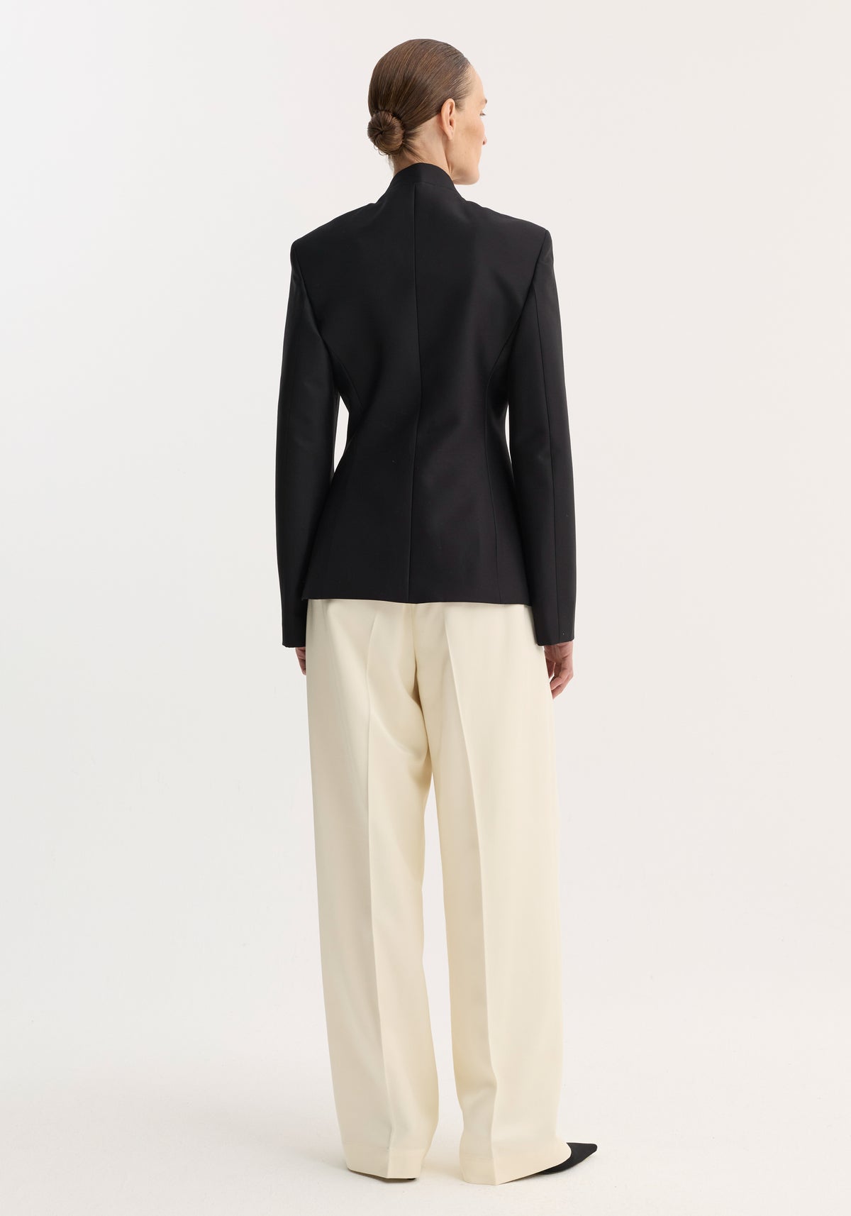 Person wearing Sculptural mandarin blazer | black, with padded shoulders and fitted waist, paired with wide-leg trousers. Hair in a neat bun, showcasing minimalist style.
