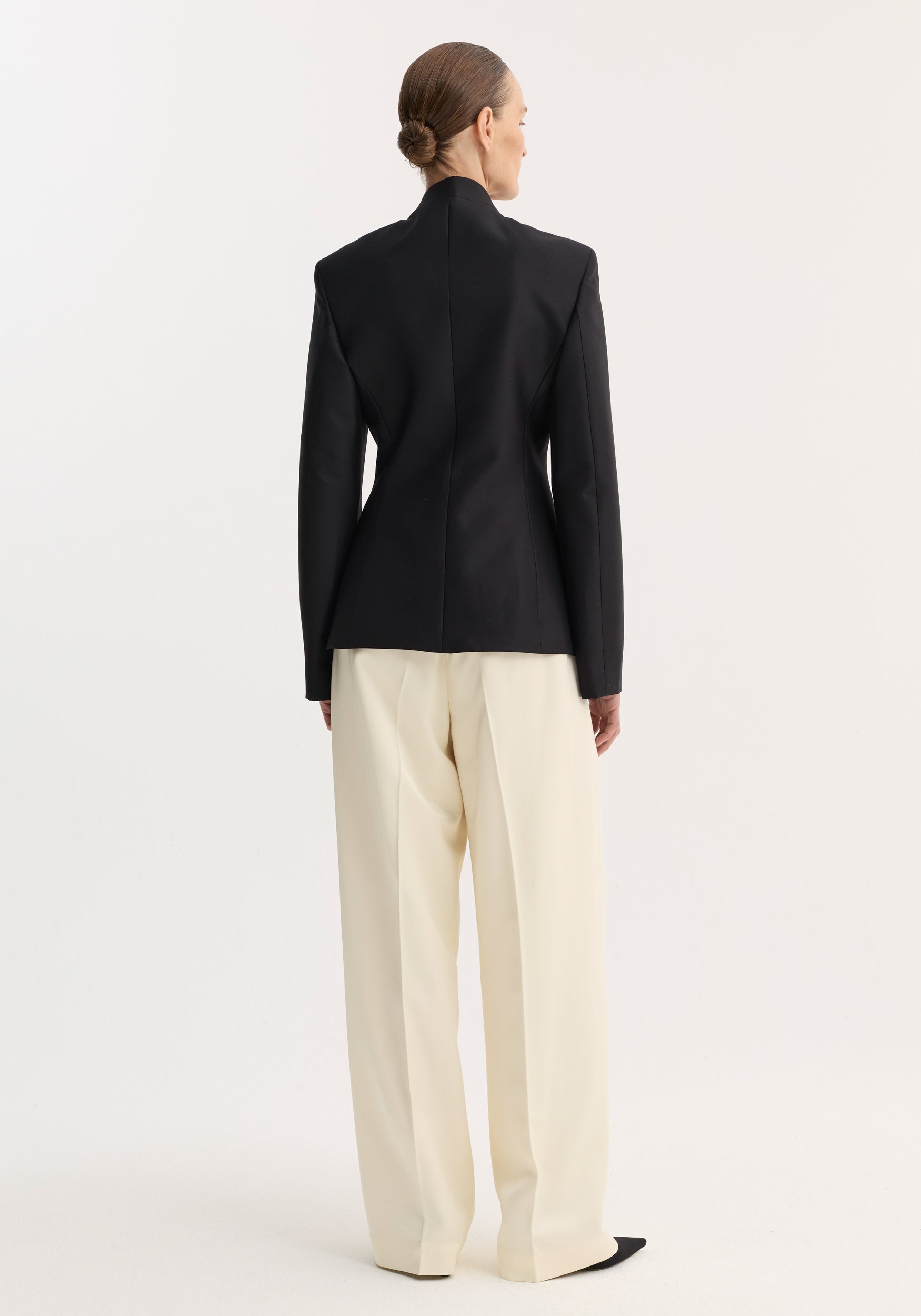 Person wearing Sculptural mandarin blazer | black, with padded shoulders and fitted waist, paired with wide-leg trousers. Hair in a neat bun, showcasing minimalist style.