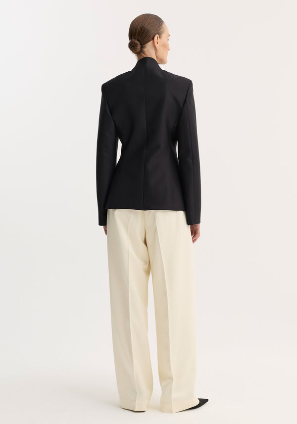 Person wearing Sculptural mandarin blazer | black, with padded shoulders and fitted waist, paired with wide-leg trousers. Hair in a neat bun, showcasing minimalist style.