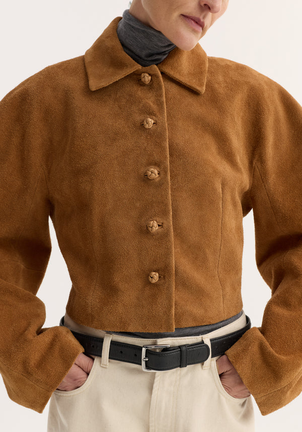 Model wearing a Rounded textured suede jacket | cognac, featuring a cropped fit with knotted buttons, over a dark turtleneck. Paired with high-waisted trousers and a black belt.