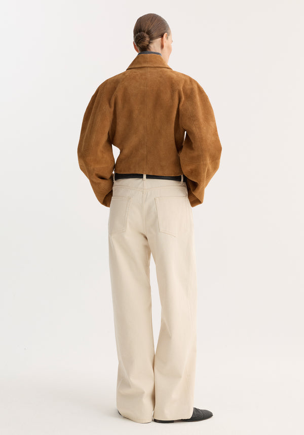 Person in a Rounded textured suede jacket | cognac, featuring puffed sleeves and knotted buttons, paired with high-waisted trousers and slip-on shoes, hair in a low bun.