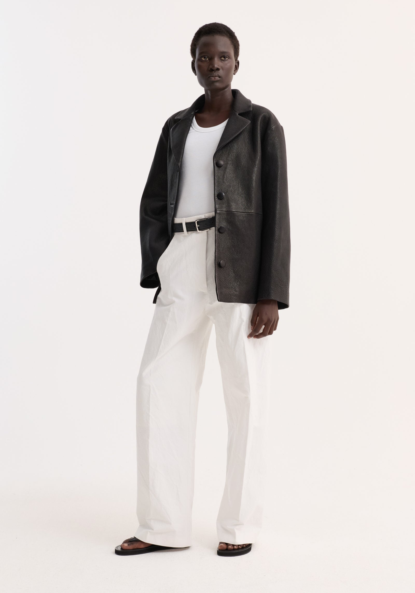 Model wearing a Leather jacket with small lapel, featuring a boxy fit, button closures, and side-seam pockets, styled over a white tank and trousers.