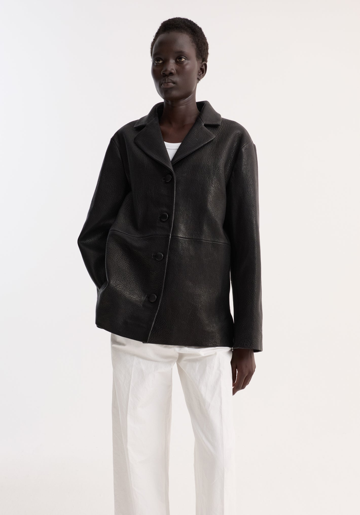 Person wearing a black leather jacket with a notched collar and buttons, styled with white trousers. The jacket has a boxy silhouette and side-seam pockets.
