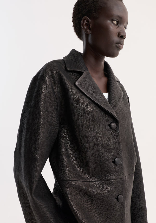 Person wearing a Leather jacket with small lapel | black, featuring a pebbled texture, notched collar, three buttons, and glimpses of a white top underneath.