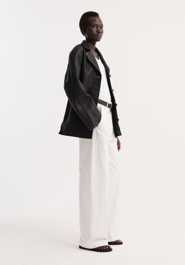 Person wearing a Leather jacket with small lapel | black, showcasing a slightly oversized, textured design, paired with white trousers and black sandals.