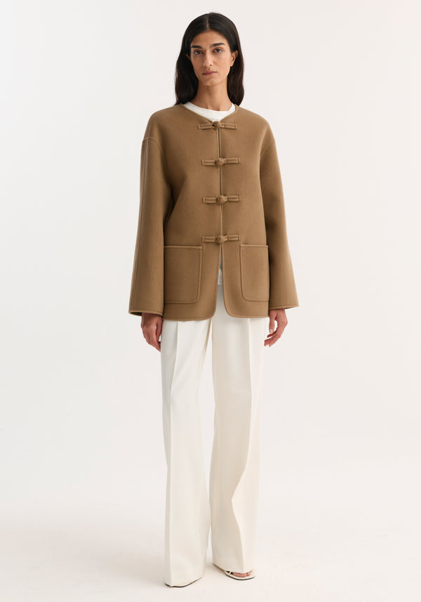 Model wearing Mandarin double-faced wool jacket in camel with pankou closure, embroidered edges, and patch pockets, paired with white trousers and shoes.