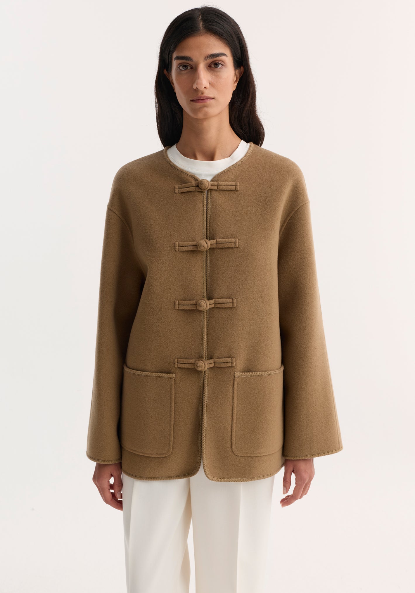 Woman wearing the Mandarin double-faced wool jacket with pankou closures, embroidered edges, patch pockets, over a white top and trousers.