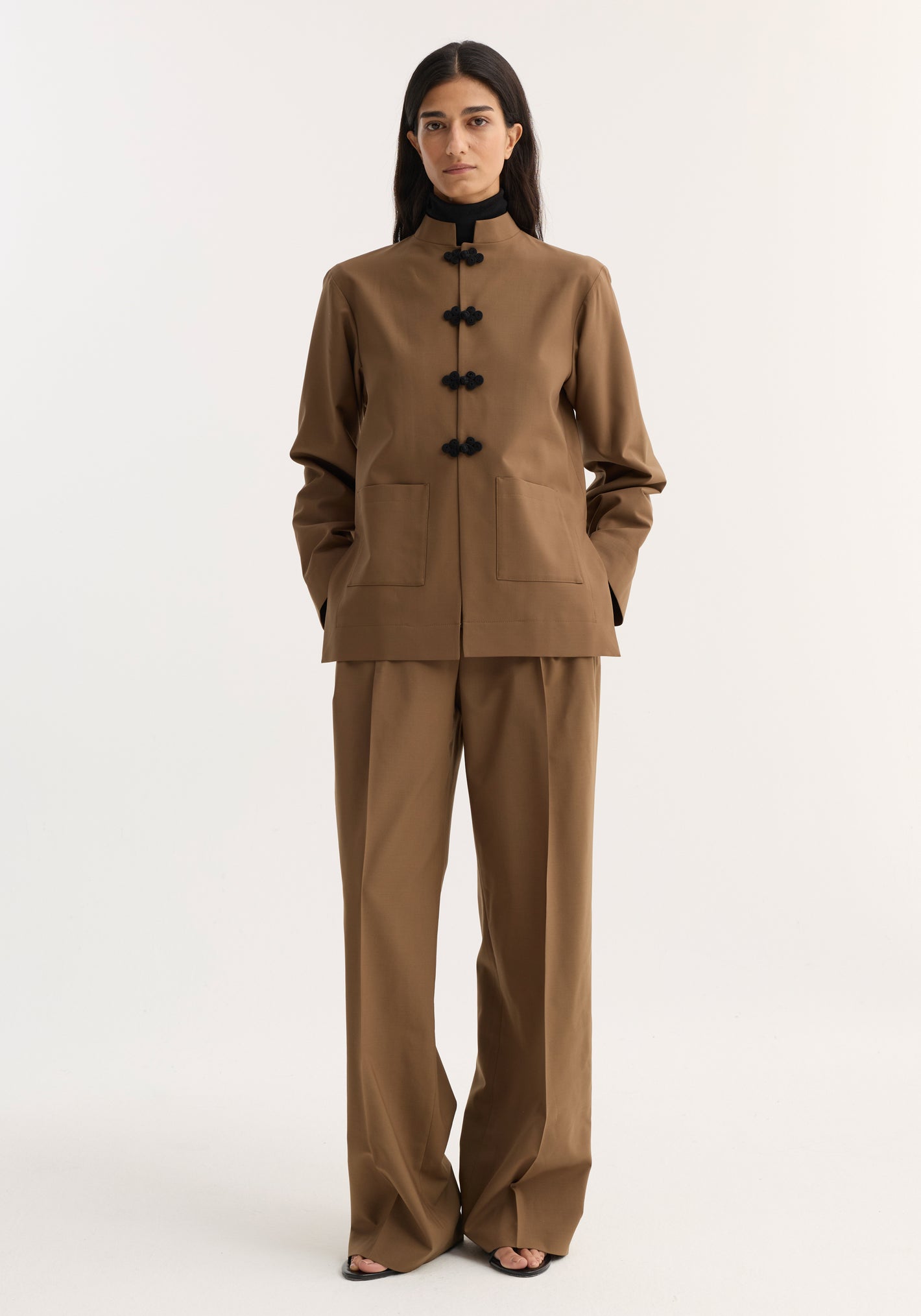 Mandarin Jacket in mocha with black frog buttons and patch pockets, worn with wide-leg trousers, black turtleneck, and open-toed sandals by a standing woman.