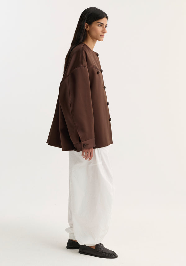Model wearing an oversized Western heavy satin jacket in espresso with leather-knotted buttons, paired with loose white trousers and black slip-on loafers.