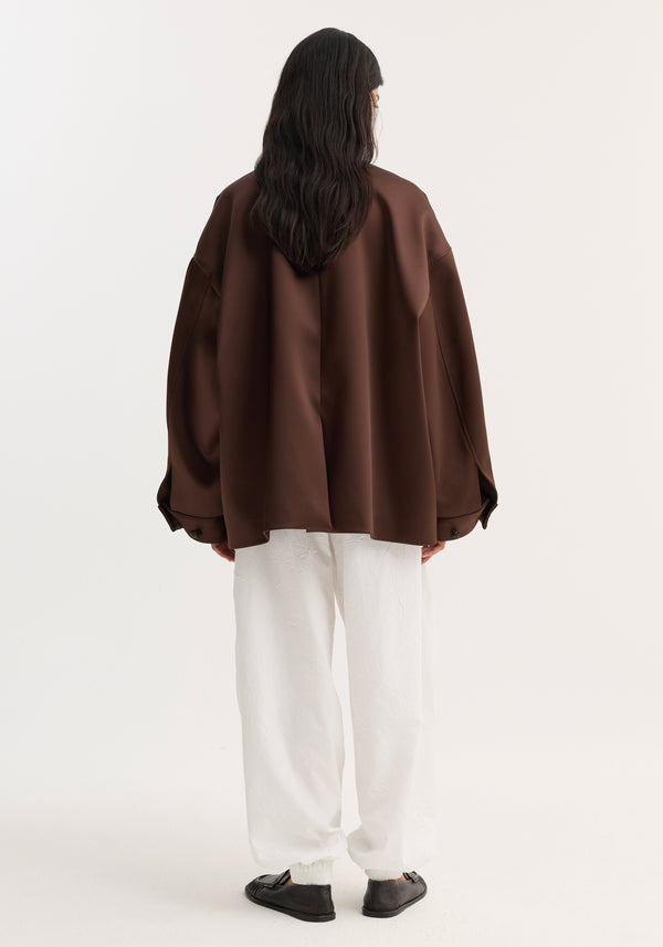 Model wears Western heavy satin jacket | espresso, featuring oversized fit, rounded sleeves, chest welt pockets, leather-knotted buttons, paired with loose white pants and black slip-on shoes.