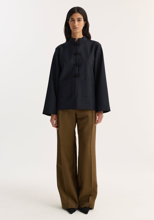 Model wearing Róhe 'Marie' Mandarin jacket with frog-style toggles and patch pockets, paired with high-waisted, wide-leg trousers.
