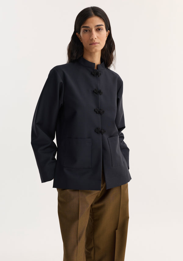 Person wearing the Róhe 'Marie' Mandarin jacket in navy with pankou closures and patch pockets, paired with loose-fit brown trousers, showcasing a minimal, tailored look.