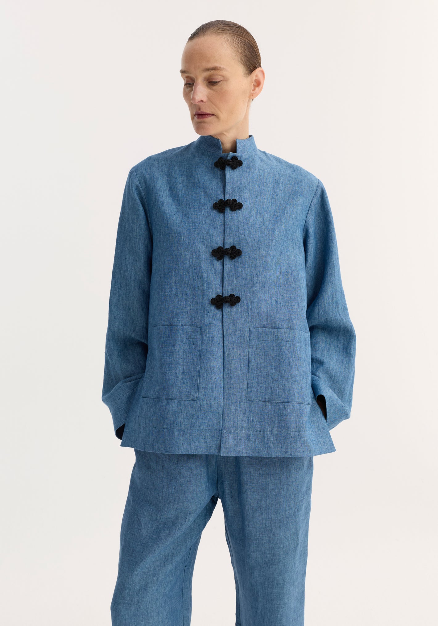 Model in an ocean blue Mandarin jacket with black frog closures and patch pockets, paired with matching straight-leg trousers, made from breathable linen.