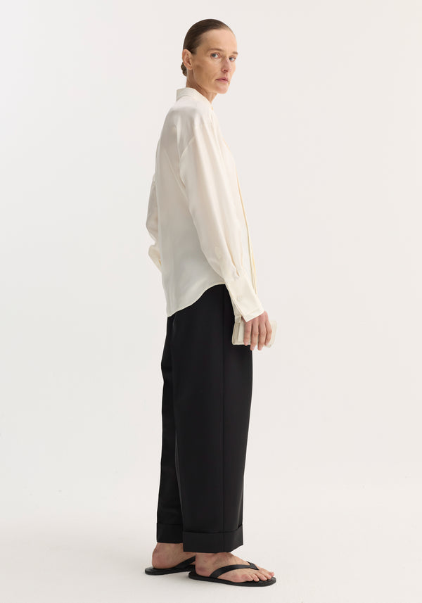 Person in profile wearing a shaped tuxedo silk shirt with a stand-up collar and pleated front, paired with wide-leg trousers and holding a clutch.