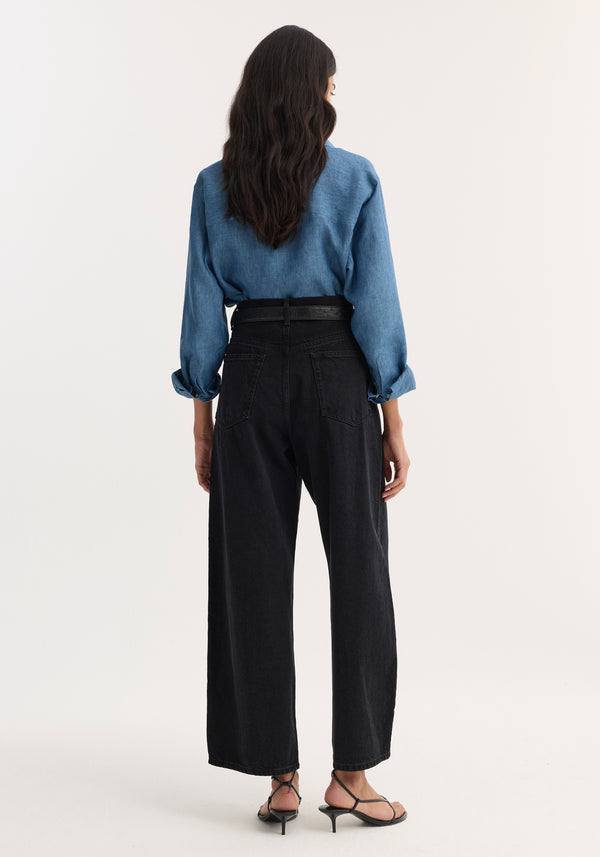 Back view of woman wearing a Classic chambray shirt in ocean blue, tucked into high-waisted black jeans, paired with strappy sandals, highlighting the shirt's relaxed fit and soft finish.