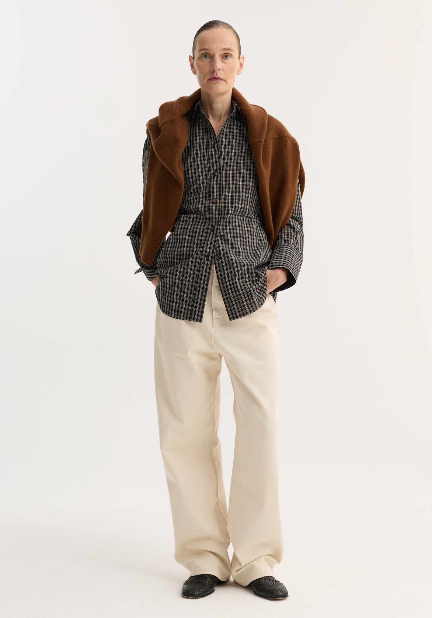 Person wearing a Checked shaped shirt in black/brown check, draped with a brown sweater, paired with cream wide-leg trousers and black slip-on shoes.