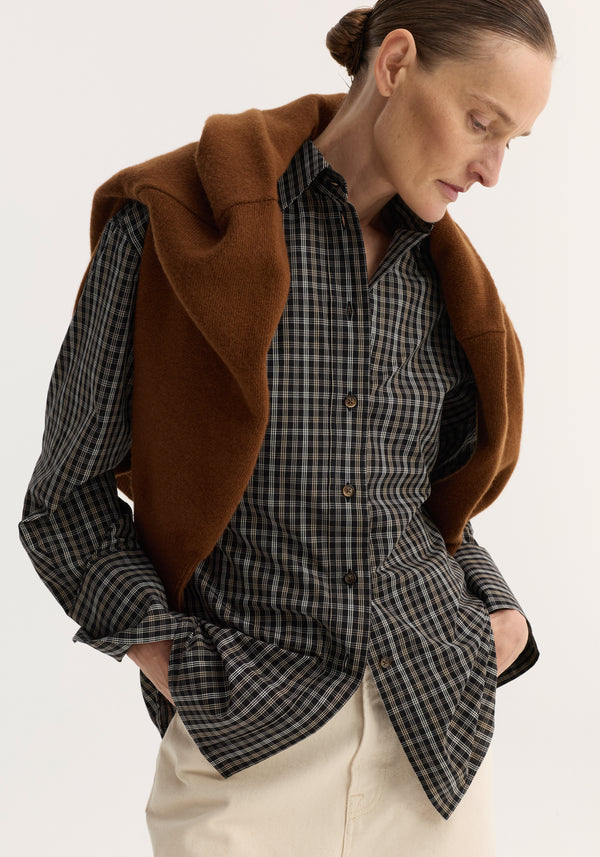 Person wearing a black-and-white checked shaped shirt with a relaxed fit and hourglass silhouette, tucked into light-beige trousers, and a brown sweater draped over shoulders.