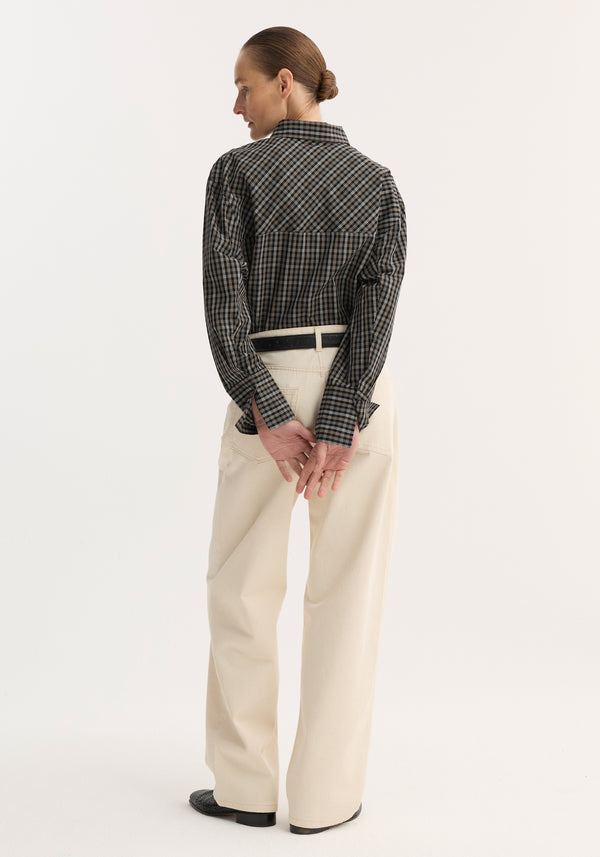 Person wearing relaxed fit, cotton checkered shaped shirt with hourglass silhouette, paired with cream trousers. Hair in bun, hands clasped behind, minimalist setting.