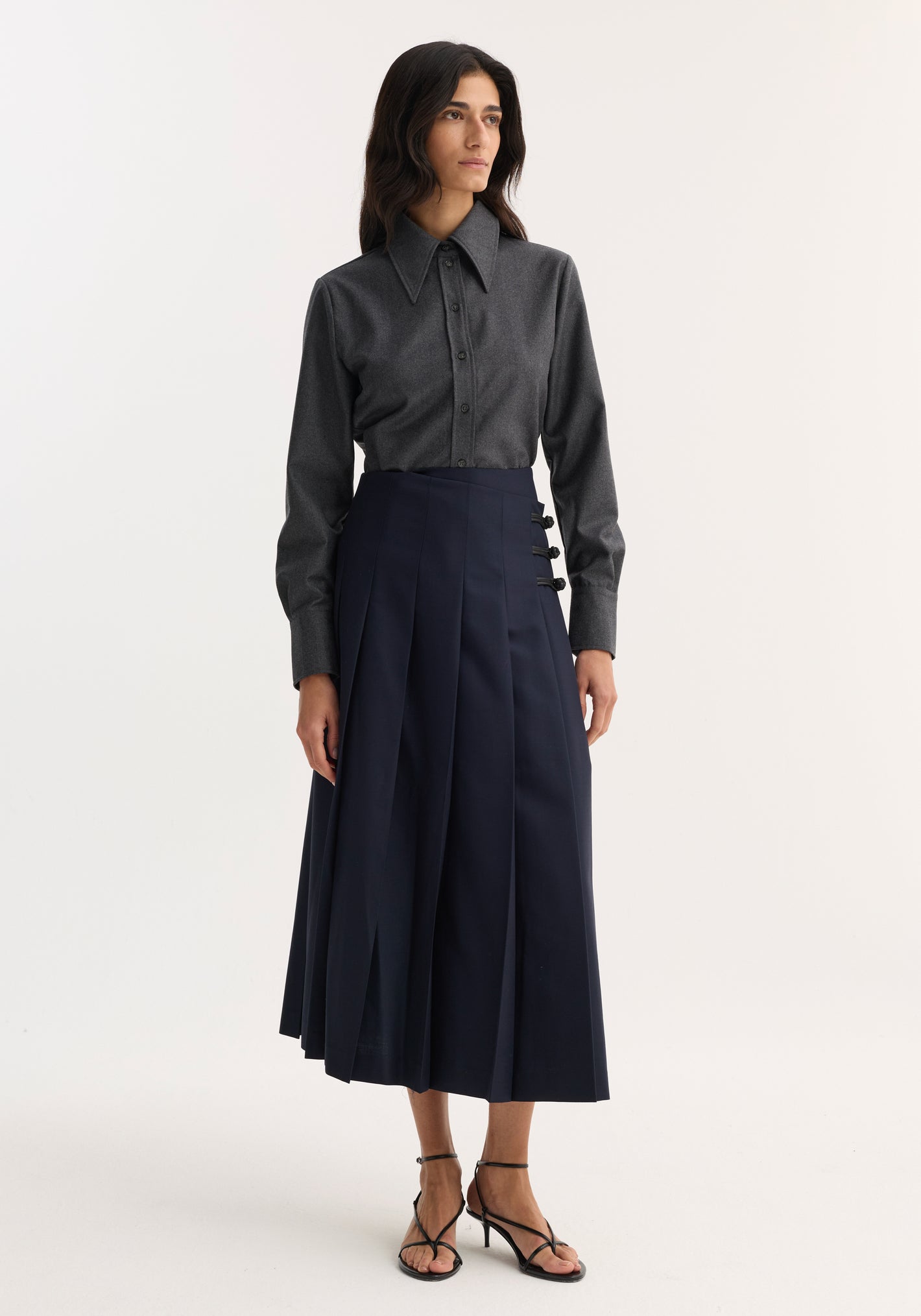 Woman in a fitted shirt, dark grey melange, with a sharp collar and button-down front, paired with a navy pleated midi skirt and strappy sandals.