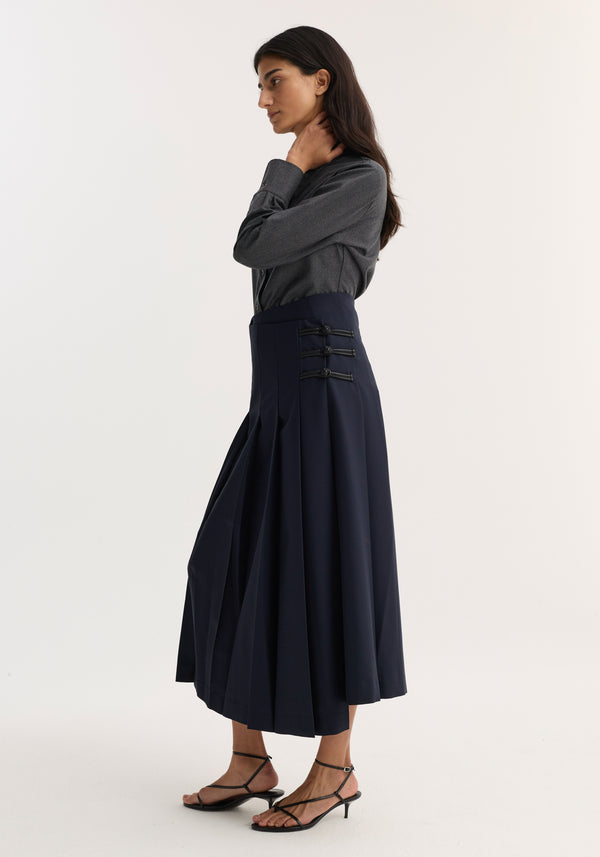 Woman in profile wearing a fitted shirt, dark grey melange, with a sharp collar, tucked into a high-waisted pleated skirt, accessorized with buckle straps.