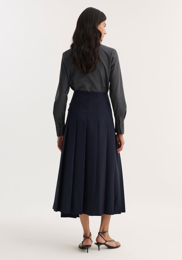 Back view of a woman in a dark navy pleated skirt, wearing a dark grey melange fitted shirt with a sharp collar and button-down front.