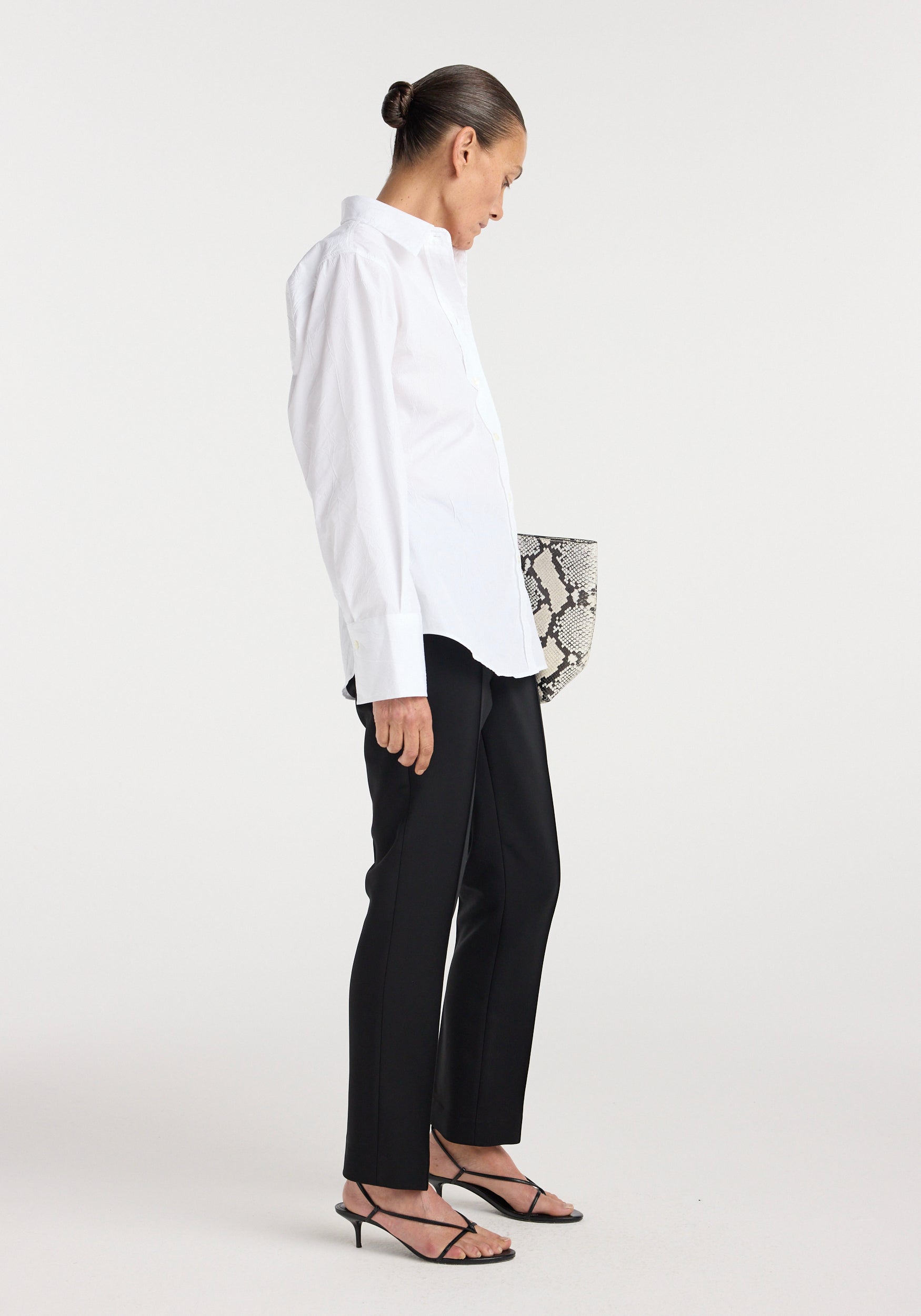 Woman wearing a shaped shirt | optic white, paired with black tailored pants and black strappy heels, holding a snakeskin-patterned clutch against a plain white background.
