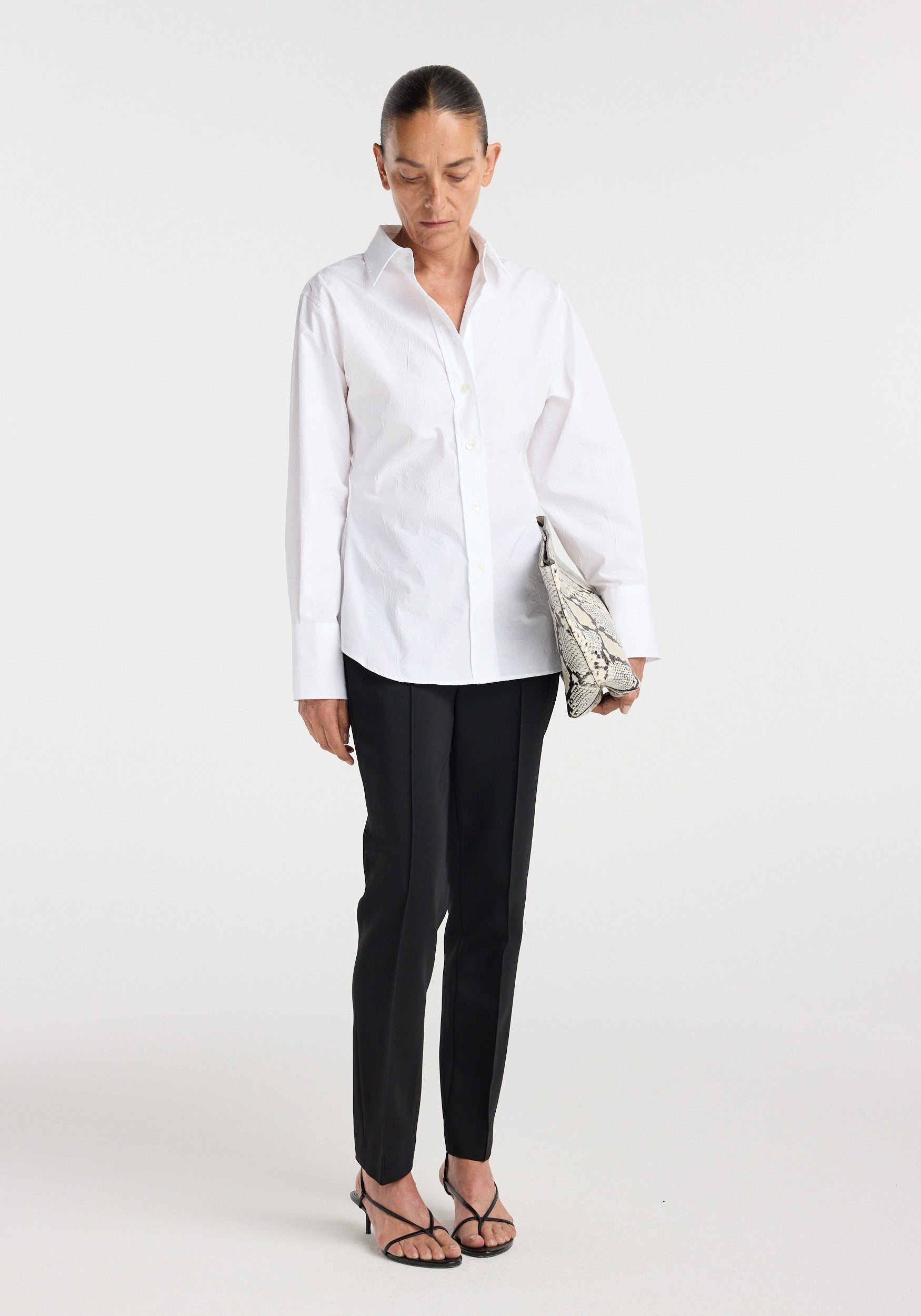 Woman wearing a crisp shaped shirt | optic white with long sleeves and button-up front, paired with black tailored pants, holding a snakeskin clutch and wearing black strappy heels.