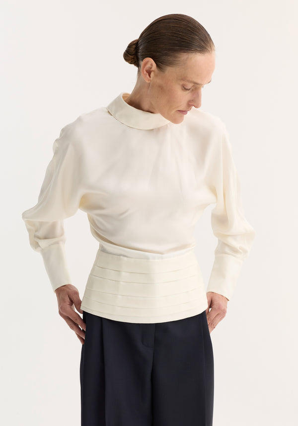 Model wearing Tuxedo silk top | cream, featuring voluminous sleeves, reversed high collar, open-back slit, and cummerbund detailing, paired with loose dark trousers.