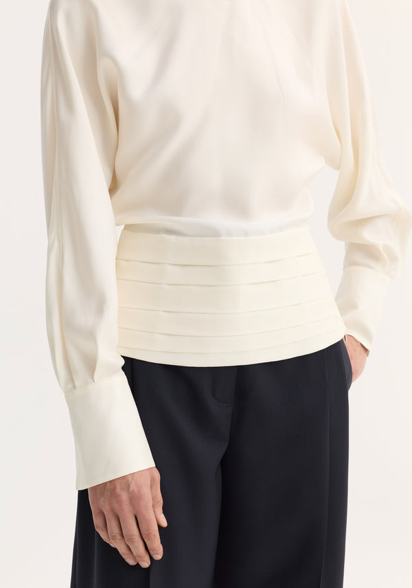 Model wearing Tuxedo silk top in cream, featuring voluminous sleeves, cummerbund waist, and pocketed dark trousers. The blouse showcases an open-back slit and reversed collar.