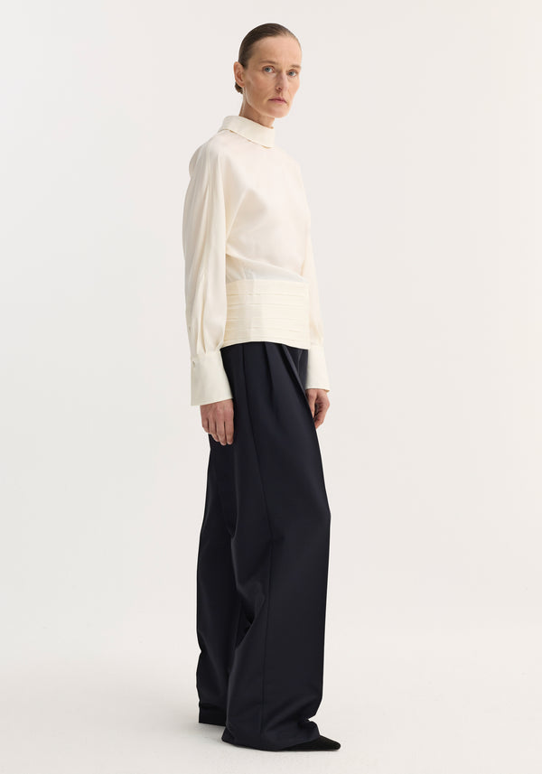 Model in profile wearing a Tuxedo silk top | cream with voluminous sleeves, high neckline, and wide-leg trousers. The blouse features an open-back slit and cummerbund detailing.