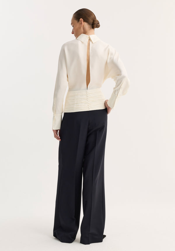 Woman in Tuxedo silk top | cream, showcasing its open-back slit, voluminous sleeves, and cummerbund detailing. Paired with high-waisted, wide-leg black trousers.