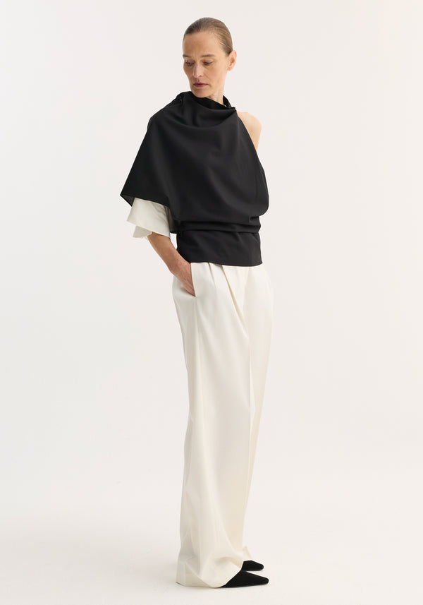 Model wearing Draped open back wool top | black, featuring an asymmetrical design with a single sleeve and high neckline, paired with high-waisted white trousers.