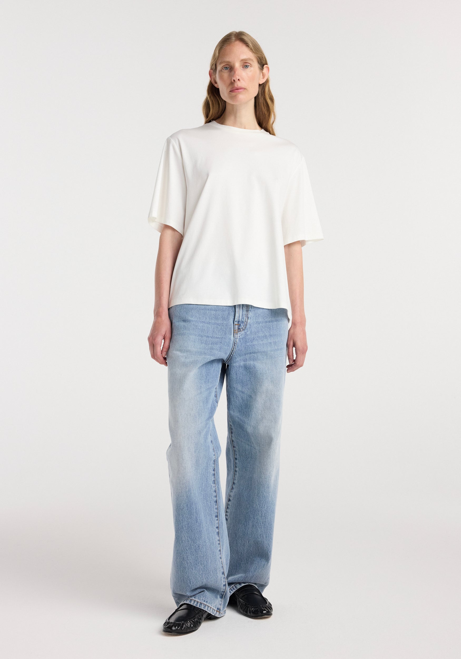 Woman wearing an oversized cotton silk t-shirt | off white paired with light blue wide-leg jeans and black shoes standing against a plain white background.
