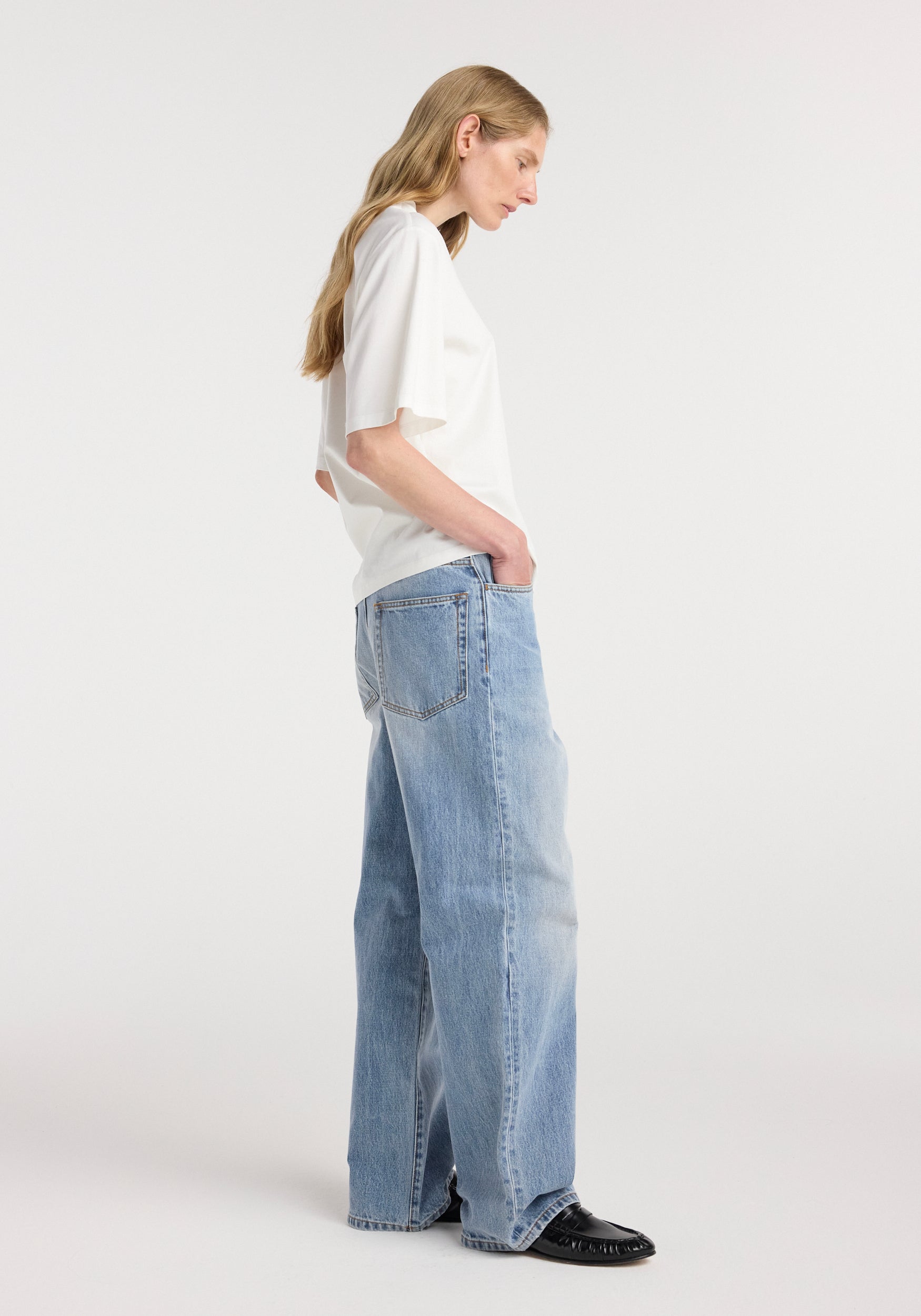 Side view of a woman wearing an oversized cotton silk t-shirt | off white paired with loose light blue denim jeans and black shoes standing against a plain white background.