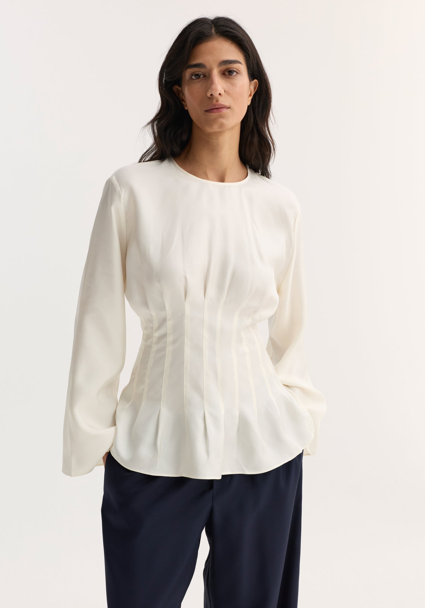 Woman wearing a Pleated silk top | cream with long sleeves, round neckline, pleated waist, and back keyhole closure, paired with wide-leg trousers.