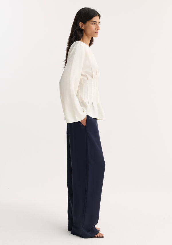 Side-profile of model wearing Pleated silk top | cream with long sleeves and subtle waist pleats, paired with navy wide-leg trousers and sandals.