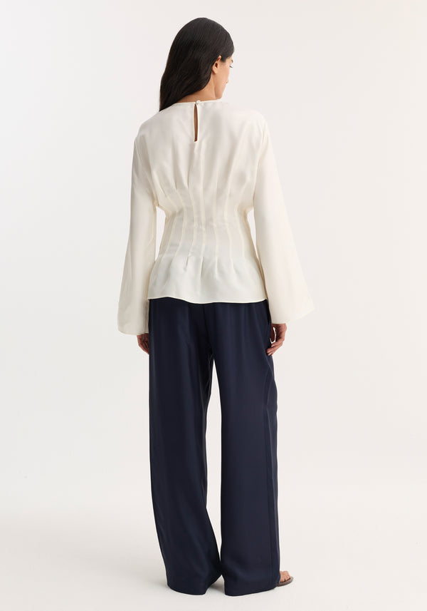Woman wearing Pleated silk top | cream, featuring long sleeves, pintucking at the waist, and a back keyhole closure, paired with navy wide-leg trousers.