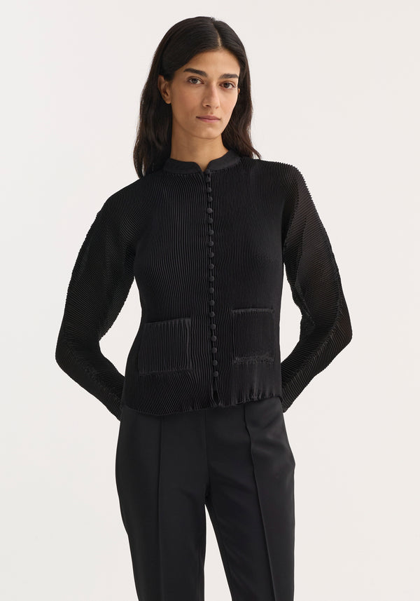 Model wearing a fitted Plissé cardigan | black with fabric-covered buttons, stand-up collar, patch pockets, and a finely ribbed texture, paired with tailored trousers.