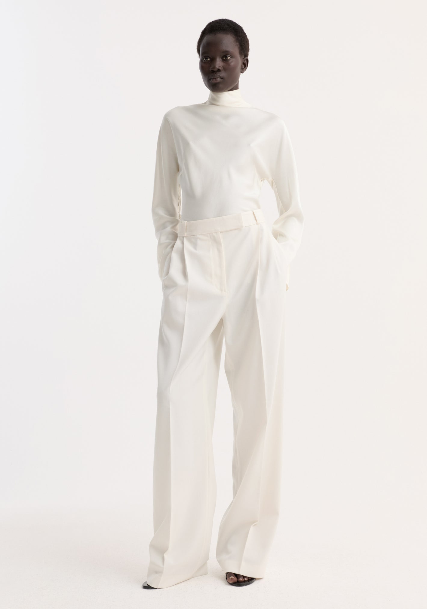Model wearing Satin waisted top | cream with high neck, long drapey sleeves, and button closures, paired with wide-leg trousers and black open-toe heels.