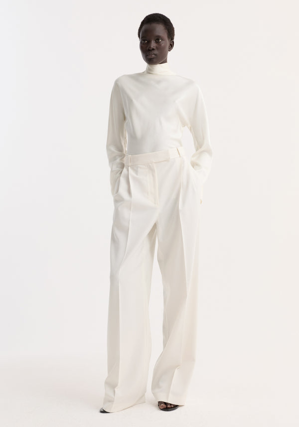 Model wearing Satin waisted top | cream with high neck, long drapey sleeves, and button closures, paired with wide-leg trousers and black open-toe heels.