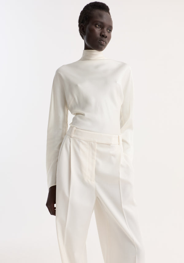 Model wears Satin waisted top | cream, showcasing a high-neck, long-sleeve satin top with button closures, paired with high-waisted tailored trousers.