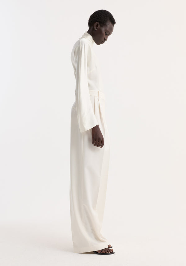 Model wearing Satin waisted top in cream, featuring fluid satin fabric, long drapey sleeves, high neckline, and delicate button closures on the back.