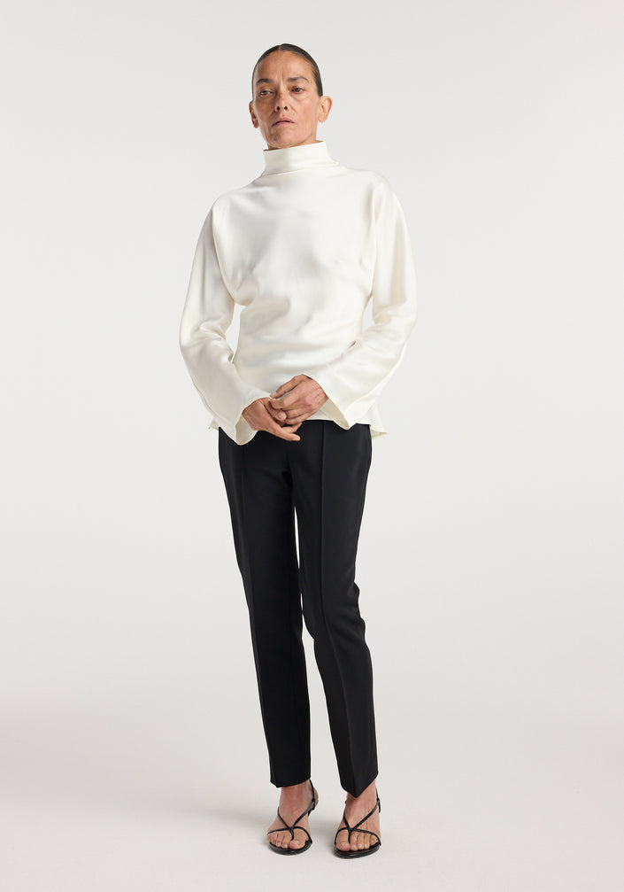 Person wearing Satin waisted top | cream in a sleek, long-sleeve design with a high neckline paired with black slim trousers and black strappy heels against a plain background.