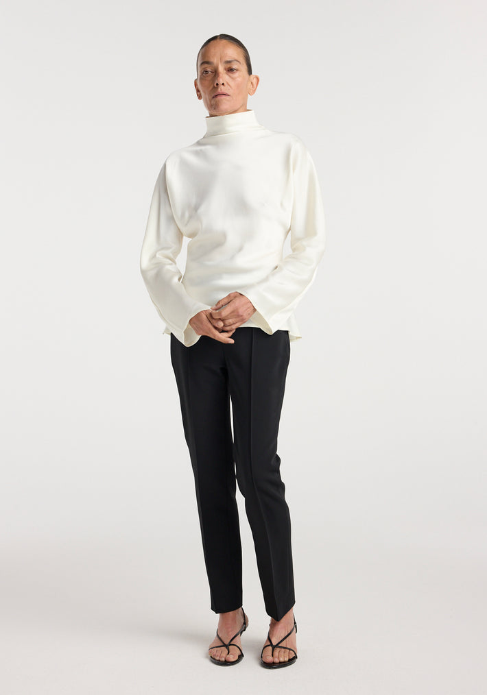 Person wearing Satin waisted top | cream in a sleek, long-sleeve design with a high neckline paired with black slim trousers and black strappy heels against a plain background.