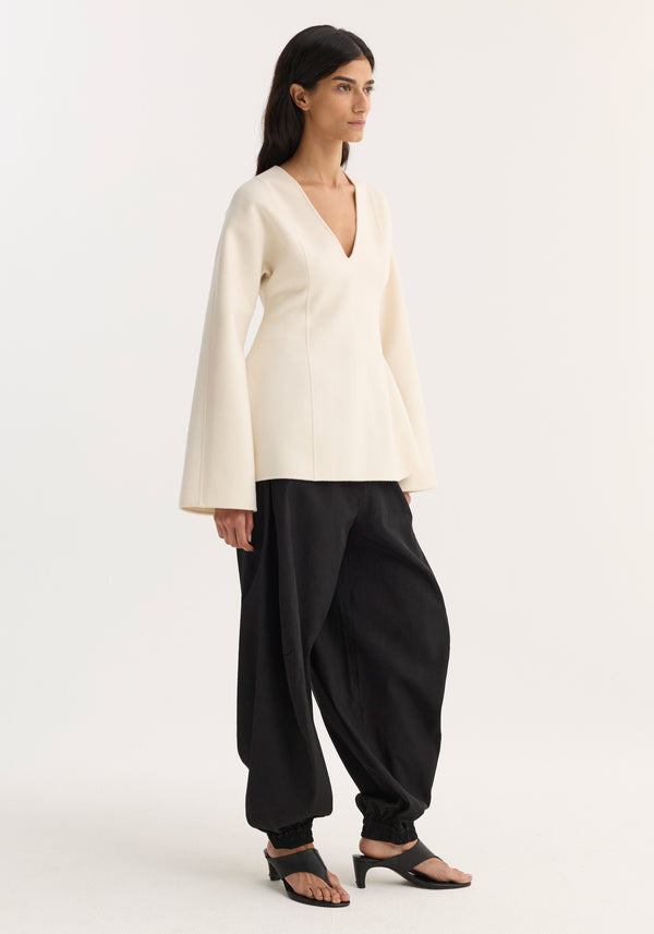 Model wearing Sculptural long sleeve top with V-neck, voluminous sleeves, and defined waist paired with black draped trousers and mid-heeled sandals.
