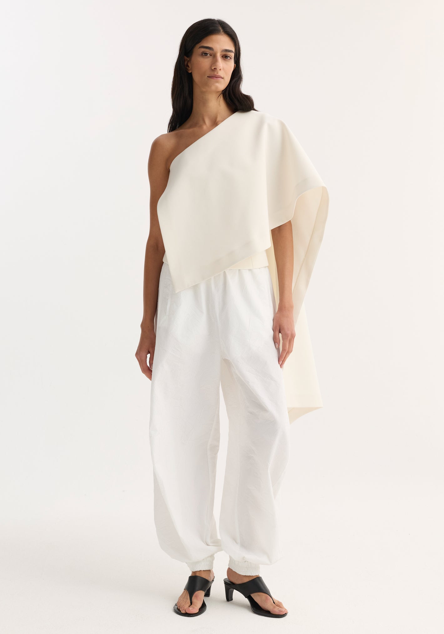 Model in cream Draped cape top with single sleeve and asymmetric hem, paired with high-waisted jogger-style pants and black open-toe heeled sandals.
